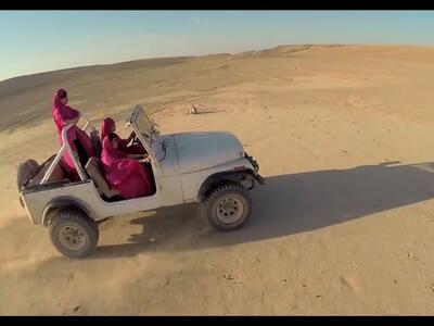 Thumbnail photo of an open-top white jeep driving across a sand dune with three women in red hijabs and gowns.