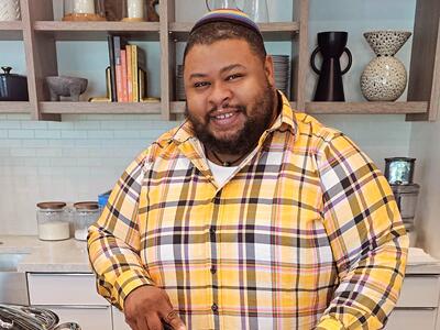A man in a yellow, black, and white plaid shirt stands at a cutting board, preparing food.