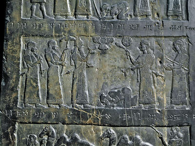 Relief of five standing figures carrying objects, and another figure prostrate in the middle of figures, surrounded by rectangular border with Akkadian inscription. 