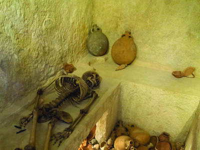 Interior room with ledges, one of which holds a skeleton, and a floor covered with pottery.