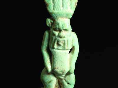 Frontal faience amulet of figure who is nude with feathered headdress and a grotesque face and beard.