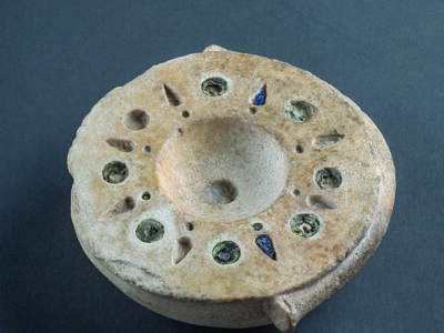 Limestone bowl with mosaic decoration around rim with alternating circular and bud-shaped inlaid blue and brown glass. 