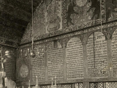 Interior of building of back wall with decorative images on top, Hebrew text in the middle, and lattice below, platform with railing in left foreground, and several podiums along wall.
