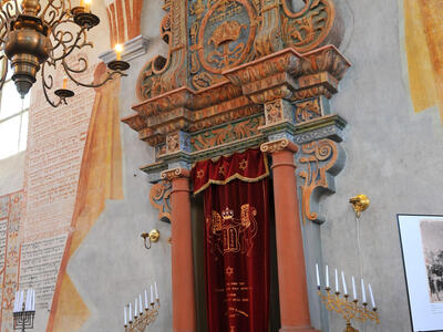 Photograph of carved doorway set in wall with curtain across it, surrounded by candelabra, Hebrew writing on wall, and chandelier in forefront. 