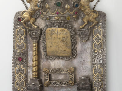 Shield with decorative border inlaid with semi-precious stones and adorned with lions and crown at top, column on left and plaque in middle with Hebrew text. 