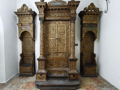 Wooden Torah ark decorated with carvings, columns, and capitals, flanked by two similarly decorated chairs. 