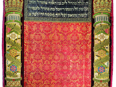 Embroidered curtain of columns with crowns on left and right sides and decorative floral motifs throughout, and Hebrew text in top center.