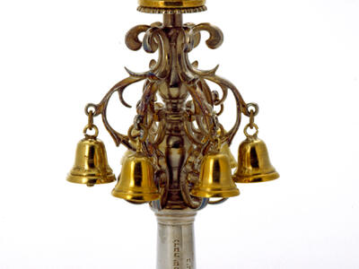 Finial with Hebrew inscription on its base, bells, and crown on top.