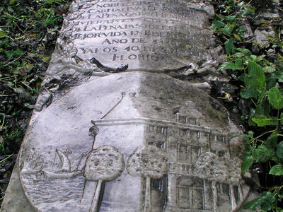 Tombstone flat on ground depicting coat of arms above Spanish inscription and relief of house surrounded by trees and next to sailboat.