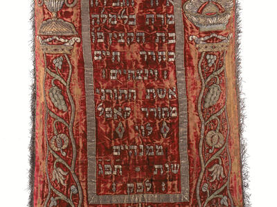 Cloth with fringe on bottom, Hebrew text on top and middle, and border of foliage. 