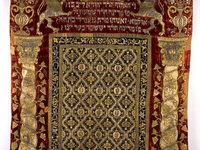 Cloth with embroidered columns on either side, pattern in the middle, and Hebrew text between two lions and crown on top.
