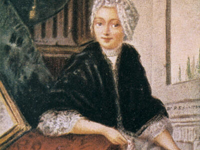 Image of seated woman in shawl with arm resting on a table with tablecloth.