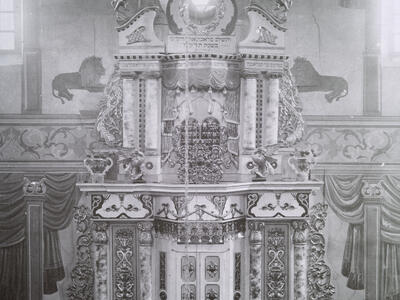 Photograph of large, ornate structure decorated with flourishes, lions, and vines, with three men in hats and beards standing in front of two closed doors at the bottom, and several other figures sitting in benches in foreground.