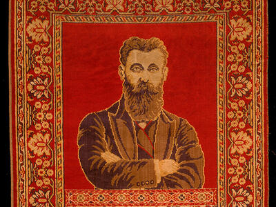 Rug depicting a bearded man with crossed arms on top half, and two columns and hanging lamp on bottom half, surrounded by decorative border.