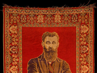 Rug depicting a bearded man with crossed arms on top half, and two columns and hanging lamp on bottom half, surrounded by decorative border.