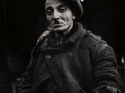 Photograph of man with mustache and hat wearing torn clothing with hands on hips, looking at camera.
