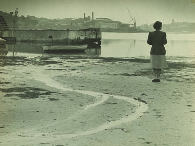 Photograph of a woman standing on a wide beach and facing away from the viewer toward a river.