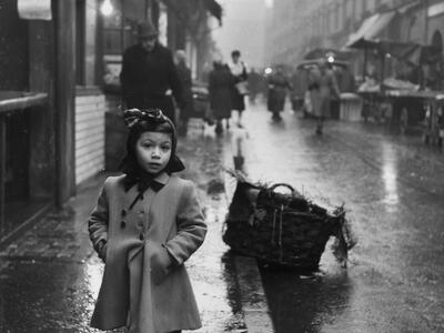 Photograph of little girl standing on rainy sidewalk of a busy city street wearing a coat and a bow facing the viewer, and basket on curb behind her.