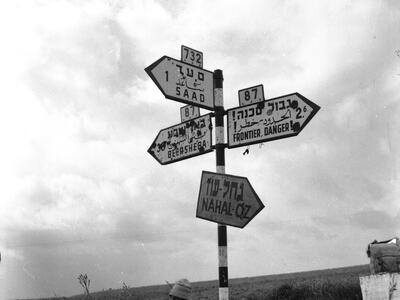 Outdoor photograph of signpost with signs pointing different directions with town names and information in Hebrew, English, and Arabic, and smiling man holding a rifle sitting on the ground beneath the sign.