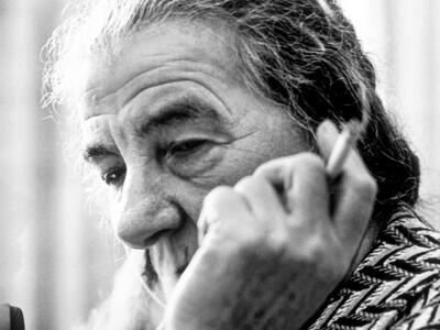 Photograph of an elderly woman smoking a cigarette, whose face is partially blocked by her hand holding the cigarette. 