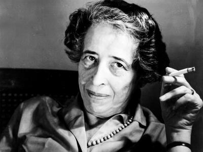 Portrait photograph of a woman looking at the camera and holding up a cigarette in her left hand.