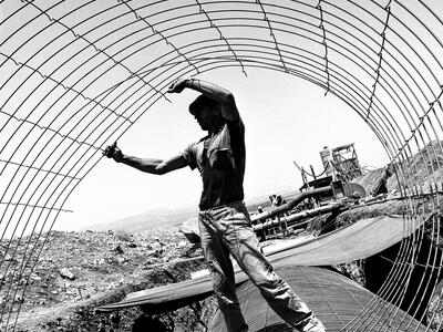 Photograph of man holding a wire cylinder and standing on a large pipe, with a construction site behind him.