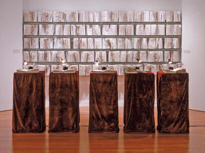 Installation featuring five podiums with an open book and reading light on top of each with open books on the wall behind. 
