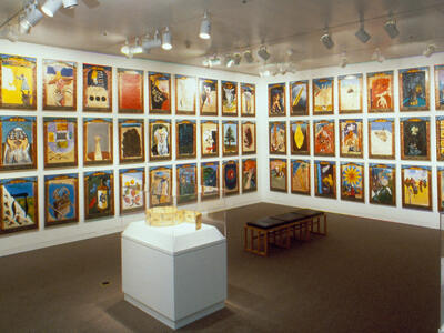 Installation photograph featuring walls of paintings and jewel case and bench in center of room.