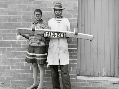 Photograph of man and woman standing and holding a car bumper in front of a brick wall.  