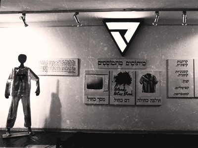 Photograph of wall with a mannequin to the left, posters with Hebrew text and an illuminated triangle on the wall, and a flag to the right.