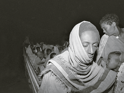 On the Way from Assembly Point Atzdemeriam to Transit Camp in Teda, Ethiopia Photograph of figure with headscarf and eyes downcast with many people behind them crowded together.