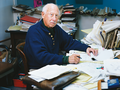 Photograph featuring older man facing viewer and seated at cluttered desk.