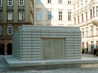 Small rectangular building made of steel and concrete with double doors on square base next to city buildings.