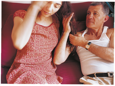 Photograph of a man sitting on a couch next to a girl and touching her hair.