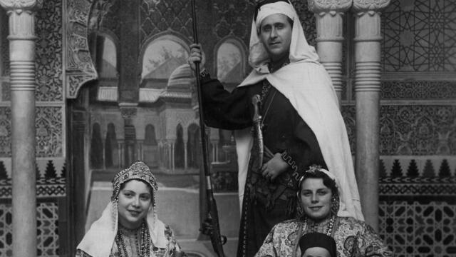 A man stands behind two seated women, one holding a baby and the other a cup and dish, all in ornate Moroccan dress.