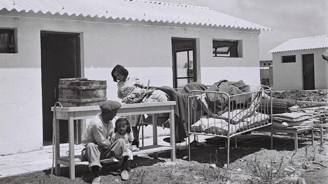 A man and two children sit outside a small building with a corrugated roof, surrounded by worn furniture and bedding.