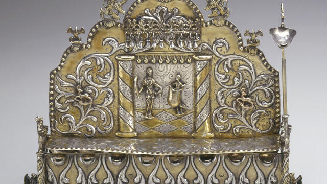Silver oil lamp with row of eight cups on bottom, and elaborate carvings of two figures and floral designs on lamp with carved birds on top. 