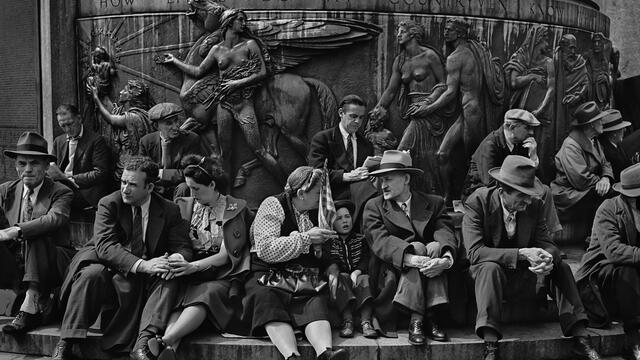 Photograph of men, women, and children seated on the steps of a large public monument with nude classical figures and a winged horse atop a circular base.