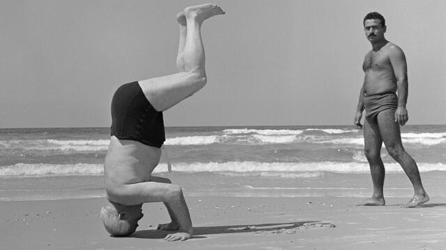 David Ben-Gurion Standing on His Head (Sharon Hotel Beach, Herzliya) Photograph of two shirtless men on the beach, one doing a headstand with legs bent facing to the right of the viewer and the other man facing the viewer on the right side of the photograph.