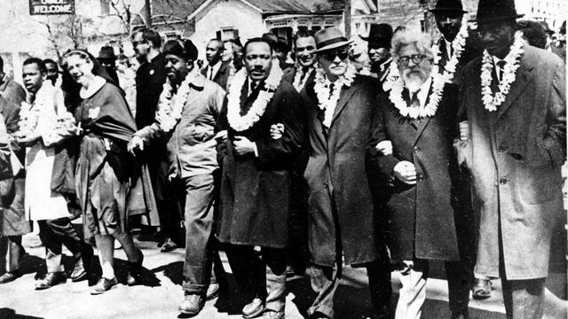Photograph of men marching down the street with arms linked wearing white double carnation leis. 